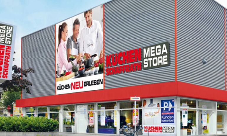Das Bild zeigt den „Küchen Schaffrath Mega Store“, ein modernes Gebäude mit einer Fassade aus grauen Metallpaneelen und roten Akzenten. Auf der linken Seite der Fassade befindet sich ein großes Werbebild mit einer Gruppe von Menschen, die gemeinsam in einer Küche kochen. Unter dem Bild steht der Schriftzug „Küchen NEU erleben“ in schwarzen und roten Buchstaben. Rechts daneben ist der Markenname „KÜCHEN SCHAFFRATH MEGA STORE“ in großen, auffälligen Buchstaben abgebildet. Im Erdgeschoss verfügt das Gebäude über große Glasfenster, die den Blick in den Verkaufsraum ermöglichen. Vor dem Gebäude befinden sich ein Fahrradständer, ein Schild für Kundenparkplätze sowie gepflegte Grünflächen. Im Hintergrund ist ein blauer Himmel mit weißen Wolken zu sehen, was eine freundliche und einladende Atmosphäre schafft.